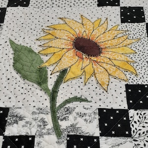 May include: A yellow and brown embroidered sunflower with green leaves on a white and black checkered background.