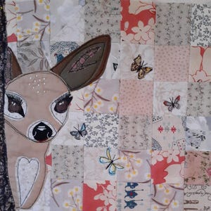 May include: A fabric wall hanging featuring a tan deer head appliqué with large dark eyes and long eyelashes, positioned next to a textured dark brown tree trunk. The background is a patchwork quilt of floral and patterned squares with butterflies.