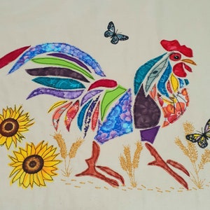 May include: A colourful patchwork rooster applique with a bright red comb and wattle. The rooster is surrounded by yellow sunflowers and green leaves. The background is a white fabric.
