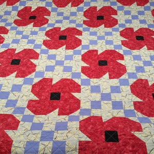 May include: A quilted blanket featuring a repeating pattern of large red poppy flowers with black centers. The flowers are set against a background of light gray fabric with a floral print and lavender squares.