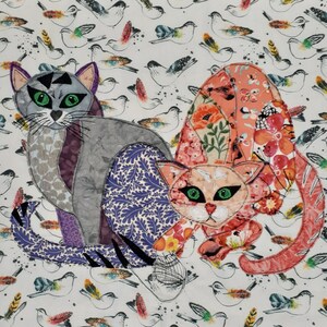 Può includere: Due gatti di stoffa colorati con motivi floreali. Il gatto a sinistra è grigio con accenti viola e neri. Il gatto a destra è rosa con accenti rossi, bianchi e verdi. I gatti sono su uno sfondo bianco con un motivo di piccoli uccelli.