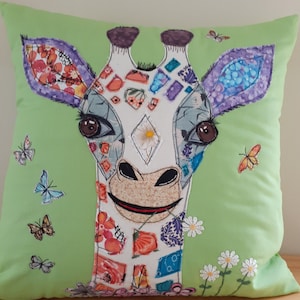 May include: A green cushion featuring a colourful patchwork giraffe design. The giraffe has a white face with brown eyes and a brown nose. The cushion is decorated with small white daisies and butterflies.