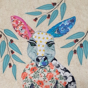 May include: A colorful patchwork fabric art piece depicting a stylized kangaroo with blue, pink, and white floral patterns. The kangaroo is surrounded by green leaves and small white flowers.