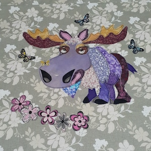 May include: A purple moose with a floral pattern on its body, with large brown antlers and big eyes. The moose is surrounded by a circle of pink and white flowers on a light grey background with a floral pattern.