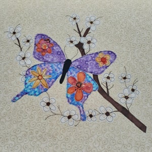 May include: A purple butterfly with colourful flower accents is appliquéd on a beige fabric background with a brown branch and white flowers.