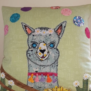 Könnte beinhalten: Ein grünes Kissen mit einem gestickten grauen Lama darauf. Das Lama trägt ein Blumenband und eine rosa-orangefarbene Quastenkette. Der Hintergrund des Kissens ist hellgrün mit einem weißen Kaktusmuster.