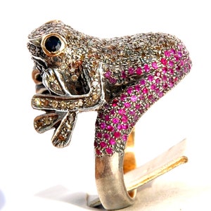 Puede incluir: Un anillo de plata con un diseño de rana. La rana está cubierta de pequeños diamantes y rubíes. El anillo está sobre un fondo blanco.