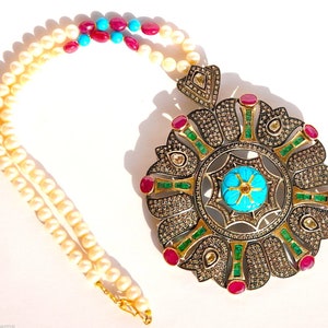 Op de afbeelding: Een zilveren ketting met een hanger met een turquoise en gouden centrum omringd door groene en rode stenen. De ketting is gemaakt van witte parels.