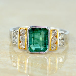 Może przedstawiać: Srebrny pierścionek z dużym szmaragdowo zielonym kamieniem szlachetnym otoczonym mniejszymi diamentami. Pierścionek ma złotą obrączkę.