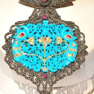 May include: A turquoise blue pendant necklace with intricate floral and bird designs. The pendant is set with black diamonds and red rubies. The necklace is made of white pearls and silver.