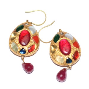 May include: A pair of gold earrings with a teardrop shape. The earrings are adorned with a cluster of colorful gemstones, including red, green, blue, and white. The gemstones are set in a gold frame and have a textured surface. The earrings have a dangling teardrop-shaped ruby gemstone.