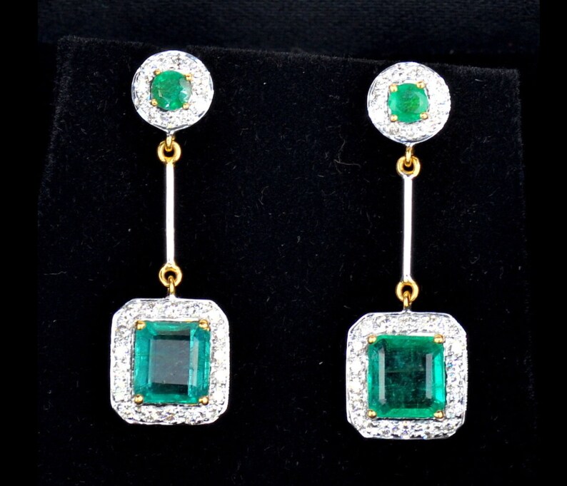 May include: A pair of emerald and diamond drop earrings. The earrings feature a square emerald surrounded by a halo of diamonds, and a smaller round emerald at the top of the earring.