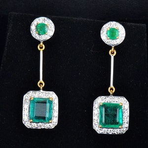 May include: A pair of emerald and diamond drop earrings. The earrings feature a square emerald surrounded by a halo of diamonds, suspended from a smaller round emerald surrounded by diamonds.