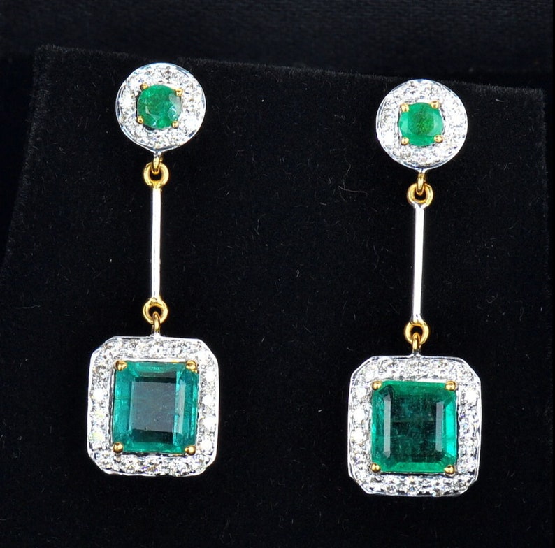May include: A pair of emerald and diamond drop earrings. The earrings feature a square emerald surrounded by a halo of diamonds, suspended from a smaller round emerald surrounded by diamonds.