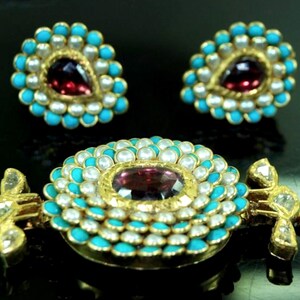 Puede incluir: Un collar de oro con un colgante redondo engastado con una gran piedra roja rodeada de perlas blancas y piedras turquesa. El colgante est&aacute; unido a una cadena de perlas blancas. El collar tambi&eacute;n incluye un par de pendientes a juego con una versi&oacute;n m&aacute;s peque&ntilde;a del colgante.