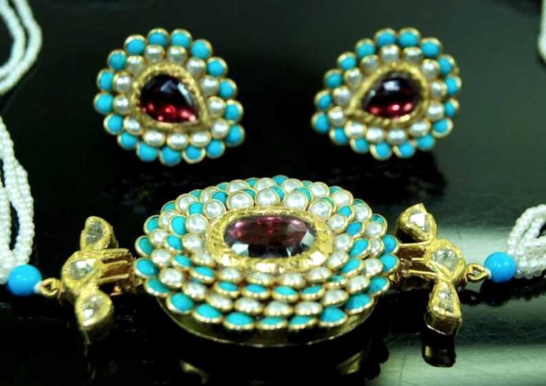 Puede incluir: Un collar de oro con un colgante redondo engastado con una gran piedra roja rodeada de perlas blancas y piedras turquesa. El colgante est&aacute; unido a una cadena de perlas blancas. El collar tambi&eacute;n incluye un par de pendientes a juego con una versi&oacute;n m&aacute;s peque&ntilde;a del colgante.