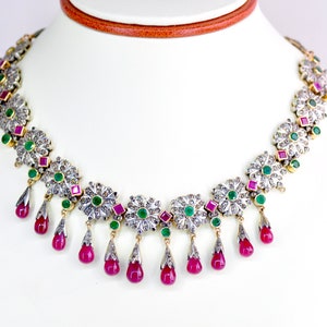 May include: A silver and gold necklace with a floral design. The necklace is adorned with small diamonds, emeralds, and rubies. The necklace has a dangling design with ruby teardrop pendants.