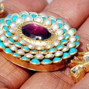 Puede incluir: Un collar con un colgante de oro redondo adornado con turquesa y perlas blancas. En el centro del colgante se encuentra una gran piedra preciosa p&uacute;rpura.