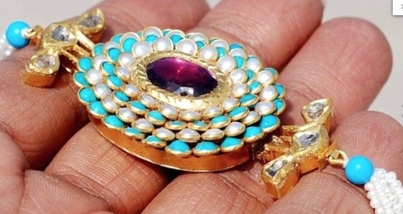 Puede incluir: Un collar con un colgante de oro redondo adornado con turquesa y perlas blancas. En el centro del colgante se encuentra una gran piedra preciosa p&uacute;rpura.