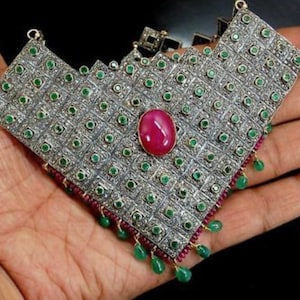 May include: A silver necklace with a large, red gemstone surrounded by smaller green gemstones. The necklace is made of a geometric pattern of squares and is decorated with small, silver beads.