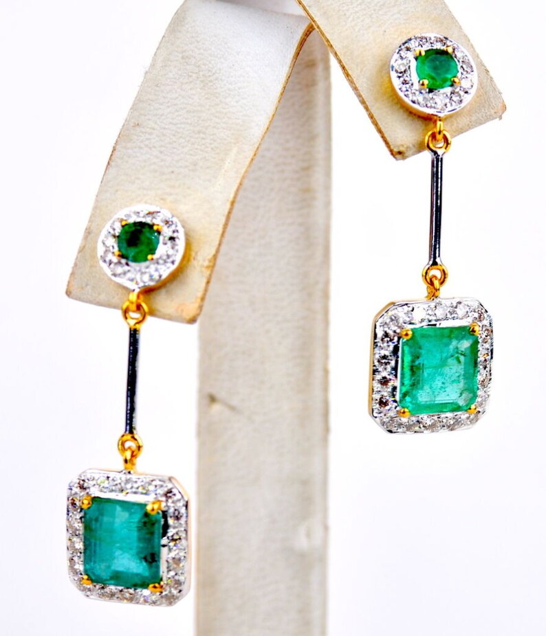 May include: A pair of gold drop earrings featuring emerald green gemstones surrounded by a halo of smaller diamonds. The earrings have a square-shaped emerald gemstone at the bottom.