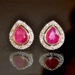 May include: A pair of gold earrings with a teardrop-shaped ruby gemstone surrounded by a halo of smaller diamonds.
