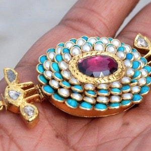 Puede incluir: Un collar de oro con un colgante circular que presenta una gran piedra roja rodeada de perlas blancas y piedras turquesa.
