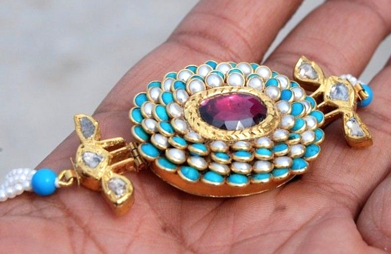Puede incluir: Un collar de oro con un colgante circular que presenta una gran piedra roja rodeada de perlas blancas y piedras turquesa.