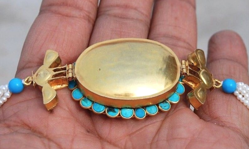 Puede incluir: Un collar de oro con un colgante ovalado de turquesa y oro. El colgante est&aacute; abierto y el interior est&aacute; vac&iacute;o. El collar est&aacute; hecho de perlas blancas y cuentas de turquesa.