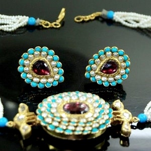 Puede incluir: Un collar de oro con un gran colgante que presenta una piedra roja rodeada de turquesa y perlas blancas. El collar est&aacute; hecho de m&uacute;ltiples hebras de perlas blancas. El conjunto tambi&eacute;n incluye un par de pendientes a juego con una versi&oacute;n m&aacute;s peque&ntilde;a del colgante.