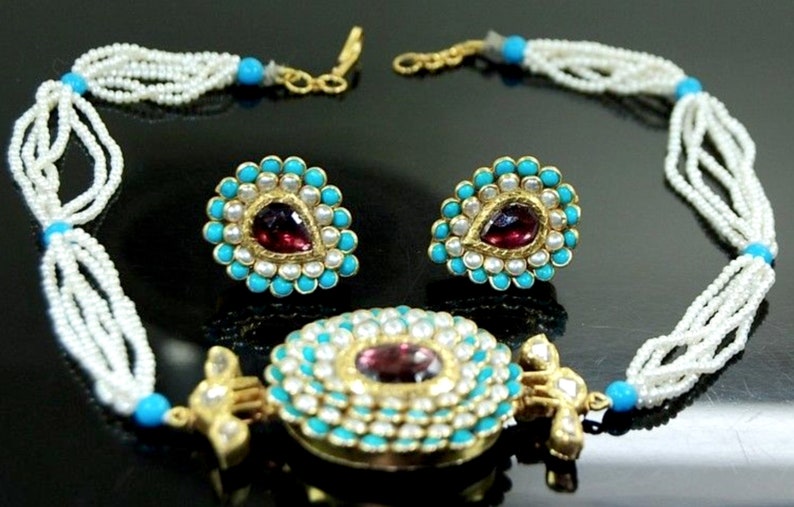 Puede incluir: Un collar de oro con un gran colgante que presenta una piedra roja rodeada de turquesa y perlas blancas. El collar est&aacute; hecho de m&uacute;ltiples hebras de perlas blancas. El conjunto tambi&eacute;n incluye un par de pendientes a juego con una versi&oacute;n m&aacute;s peque&ntilde;a del colgante.