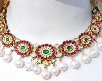 May include: A gold necklace with a floral design featuring red and green gemstones. The necklace has a row of pearls hanging from it.
