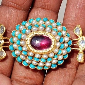 Puede incluir: Un collar de oro con un colgante grande de forma ovalada de piedra rosa rodeado de turquesa y perlas blancas. El colgante est&aacute; unido a una cadena de oro con dos colgantes m&aacute;s peque&ntilde;os en forma de diamante a cada lado.