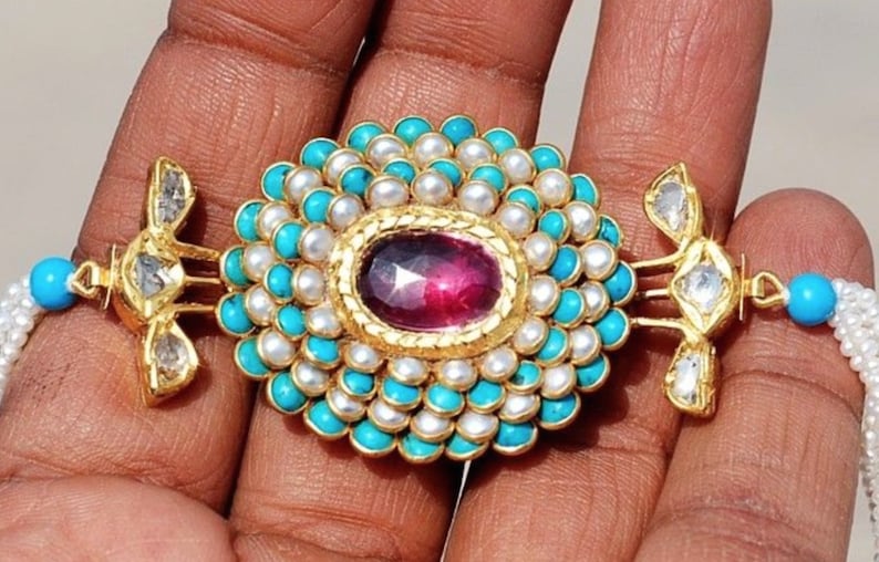 Puede incluir: Un collar de oro con un colgante grande de forma ovalada de piedra rosa rodeado de turquesa y perlas blancas. El colgante est&aacute; unido a una cadena de oro con dos colgantes m&aacute;s peque&ntilde;os en forma de diamante a cada lado.