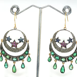 Op de afbeelding: Zilveren en gouden oorbellen met een halve maan ontwerp, versierd met drie sterren met blauwe en roze edelstenen. De oorbellen hebben druppelvormige hangers in smaragdgroen.