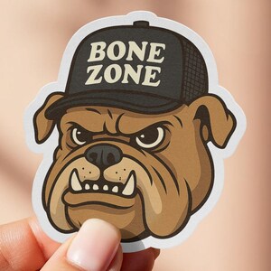 May include: A sticker featuring a cartoon bulldog wearing a black cap with the words "BONE ZONE" in white. The bulldog is brown with an angry expression and white teeth.