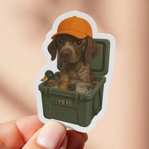 Puede incluir: Una pegatina con un perro de caza con una gorra naranja, sentado dentro de una nevera verde. La nevera contiene señuelos de pato. La pegatina tiene un borde blanco y la sostiene una mano.