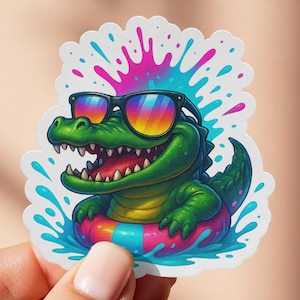 May include: A sticker featuring a cartoon alligator wearing sunglasses and floating in a colorful inner tube. The alligator is green with a yellow belly and has a wide grin. The sticker has a white border and a splash of blue and pink.