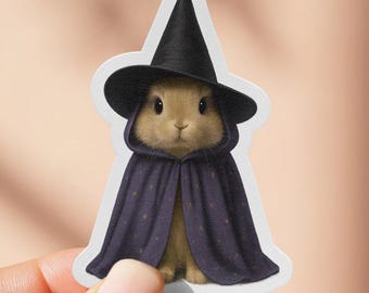 Puede incluir: Una pegatina con un conejo marrón que lleva un sombrero de bruja negro y una capa morada oscura con lunares blancos. El conejo está centrado en una pegatina blanca con borde redondeado, sostenida por una mano.