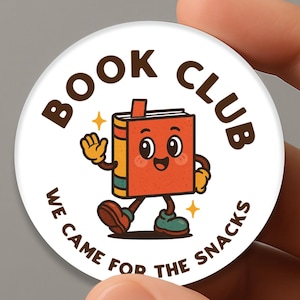 Könnte beinhalten: Ein weißer, runder Button mit den Worten "BOOK CLUB" und "WE CAME FOR THE SNACKS" in schwarzer, geschwungener Schrift. Der Button zeigt eine Cartoon-Buchfigur mit lächelndem Gesicht, winkender Hand und laufenden Beinen.