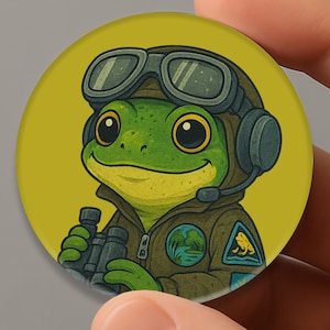 May include: A round, yellow button featuring a cartoon frog in a pilot's uniform, complete with goggles and a headset. The frog is holding binoculars and has a patch on its jacket. The button is held in a hand.
