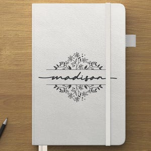 Può includere: Un taccuino bianco con il nome "madison" in corsivo, incorniciato da un disegno floreale. Il taccuino ha un elastico abbinato e un segnalibro a nastro. Una penna, una matita e uno smartphone sono anche sulla superficie di legno.