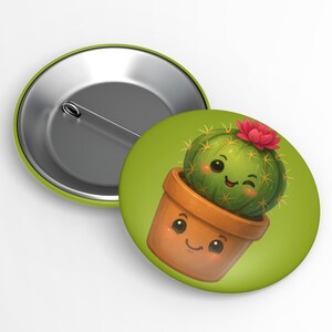 May include: A green button with a cartoon cactus in a brown pot. The cactus has a pink flower and is winking. The pot and cactus have smiling faces. The button has a green border and a metal pin back.
