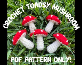 May include: Five crocheted toadstool mushrooms with white stems and red caps with white dots. The mushrooms are arranged on a bed of green grass. The text "CROCHET TOADSY MUSHROOM PDF PATTERN ONLY!" is written in white letters on a black background.