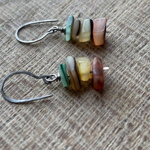 Puede incluir: Un par de pendientes hechos a mano con ganchos plateados y una disposición vertical de cuentas rectangulares de colores. Las cuentas son en tonos verdes, crema, amarillos, naranjas y marrones. Los pendientes se muestran sobre una superficie de madera texturizada.