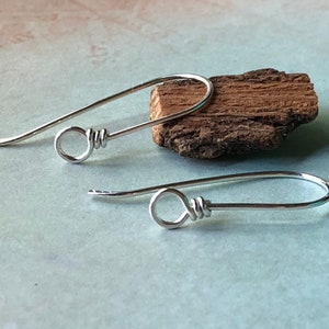 May include: Two silver-colored earring hooks with a looped design. The hooks are curved and have a simple, elegant style. The earrings are displayed on a piece of brown wood against a light blue background.