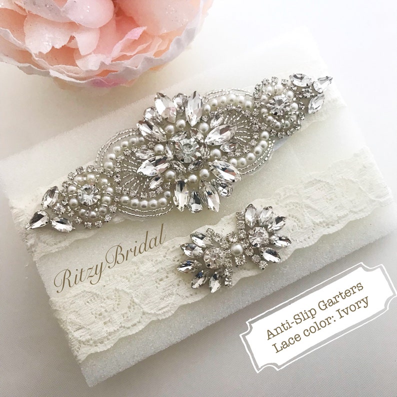 A stunning wedding garter set, designed for the bride on her special day. The main garter features a beautiful ivory lace band adorned with intricate rhinestone embellishments, adding a touch of sparkle and sophistication