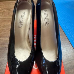 May include: Pair of black patent leather Bandolino pumps with pointed toes. The shoes have a glossy finish and a low heel. The interior is a light beige color, and the words "BANDOLINO MADE IN ITALY" are printed inside each shoe.
