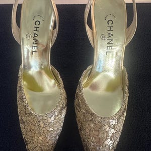 Puede incluir: Un par de tacones dorados con tiras traseras. Los zapatos presentan una puntera afilada y un delicado patrón de encaje texturizado. La plantilla está estampada con la marca "CHANEL" y "MADE IN ITALY".