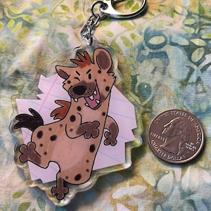 May include: A clear acrylic keychain with a cartoon hyena character, brown spots, a red mane, and a wide grin. The keychain has a silver-coloured clasp and a small metal ring. A U.S. quarter is next to the keychain.